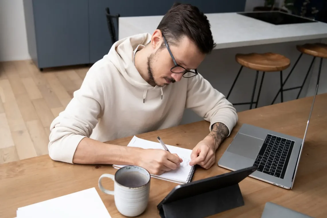 Preparação para o Concurso PF – Estudo e Foco Homem estudando com caderno, tablet e laptop sobre uma mesa de madeira, representando a preparação para concursos públicos