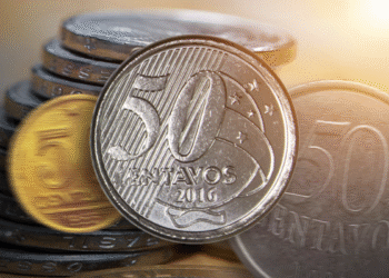Moeda brasileira de 50 centavos em destaque sobre uma pilha de moedas, com iluminação quente ao fundo.