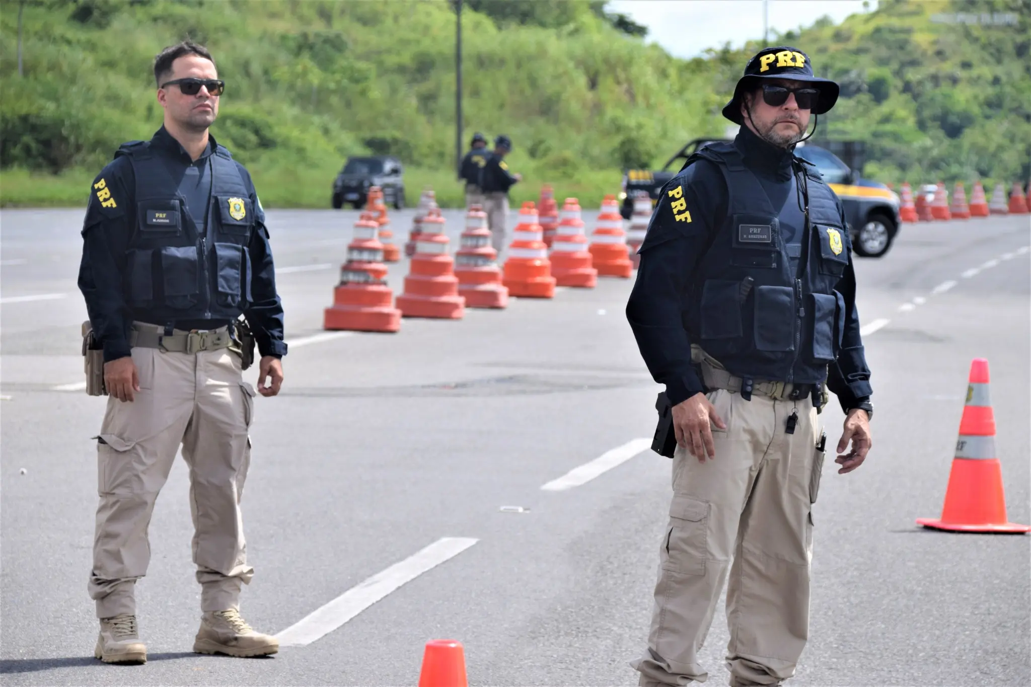 Policiais rodoviários federais em operação de fiscalização com cones de trânsito ao fundo.