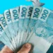 Notas de 100 reais em mãos, simbolizando indícios de riqueza ou compensações financeiras.