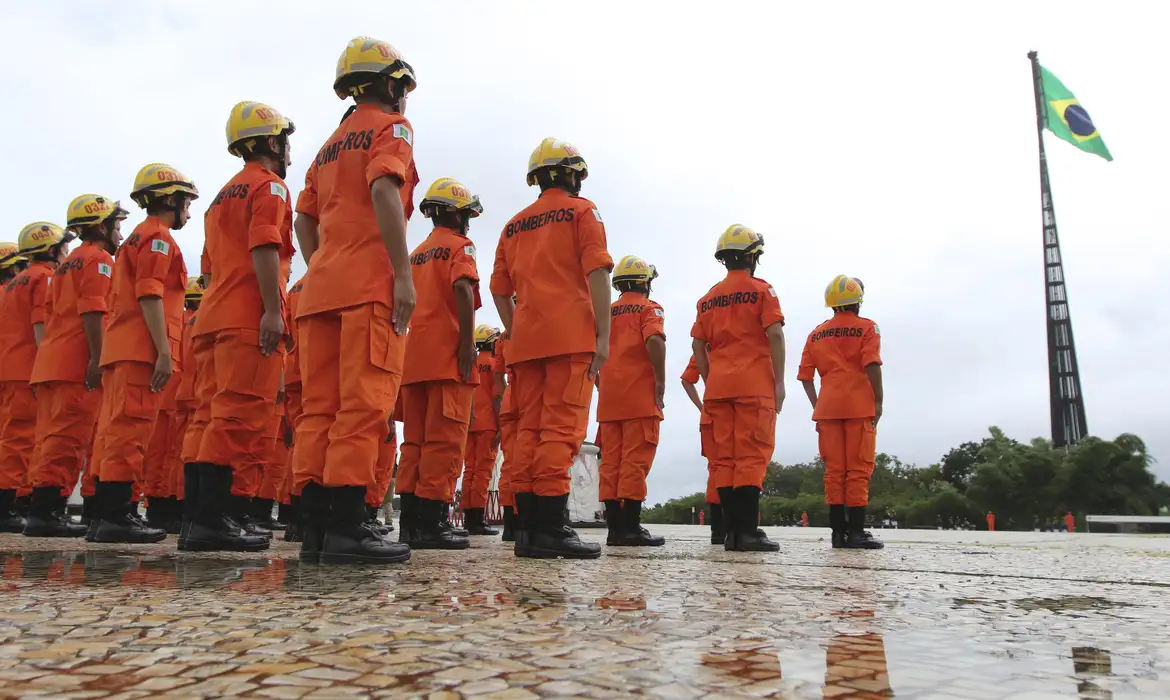 Concurso Bombeiros Bombeiros alinhados de uniforme laranja, capacetes amarelos e bandeira do Brasil ao fundo.