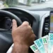 Motorista de Uber segurando o volante enquanto dirige, com notas de dinheiro em destaque