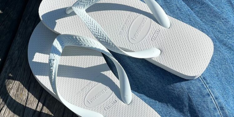 Um par de sandálias Havaianas brancas, modelo Slim, sobre uma calça jeans em uma superfície de madeira.