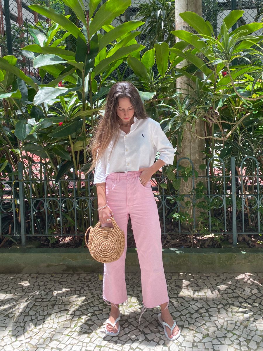 Mulher jovem, com blusa branca e calça rosa, usando Havaianas brancas e segurando uma bolsa de palha, em um cenário de jardim.