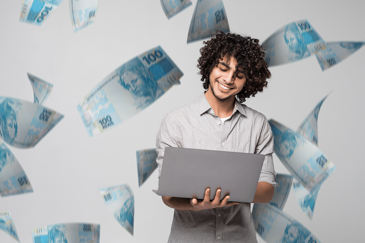 Jovem sorrindo usando laptop com notas de 100 reais voando ao fundo.