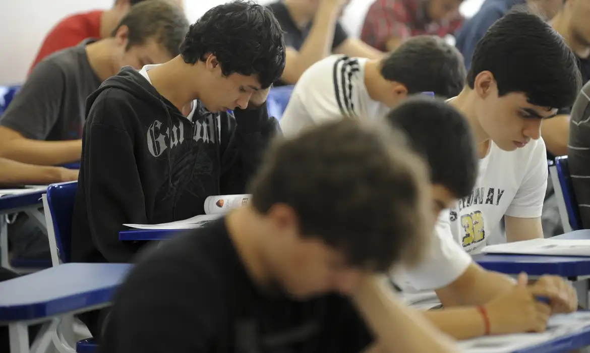 Estudantes concentrados realizando prova escrita em carteiras escolares.