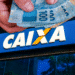 Pessoa segurando cédulas de real próximo à fachada da Caixa Econômica