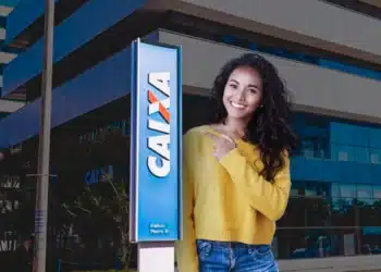 Jovem sorrindo em frente a uma agência da CAIXA, representando o anúncio do calendário de pagamentos de R$ 200 do Pé-de-Meia de setembro.