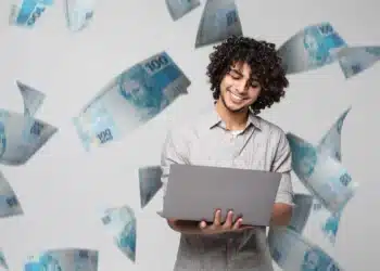Homem sorrindo com laptop enquanto notas de dinheiro caem ao seu redor, simbolizando organização financeira e tranquilidade para o fim de ano.