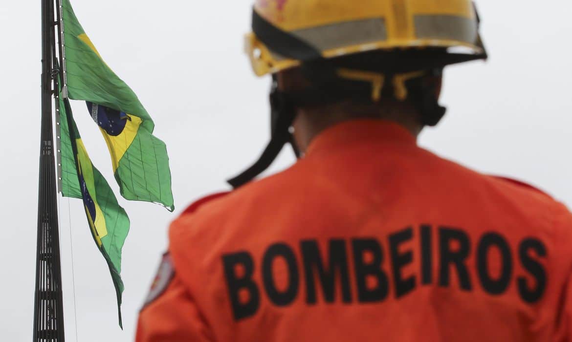 Bombeiro com capacete e uniforme laranja com a palavra Bombeiros, bandeiras do Brasil ao fundo