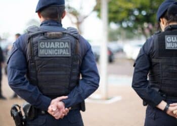Dois guardas municipais de costas em uniforme azul escuro, com colete à prova de balas e mãos entrelaçadas atrás.