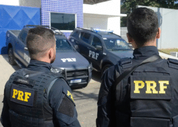 Dois policiais da PRF de costas usando coletes táticos em frente a viaturas oficiais da Polícia Rodoviária Federal