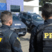 Dois policiais da PRF de costas usando coletes táticos em frente a viaturas oficiais da Polícia Rodoviária Federal