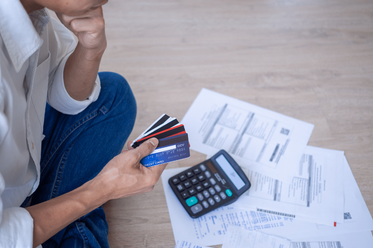 Pessoa segurando vários cartões de crédito, analisando contas com uma calculadora ao lado.