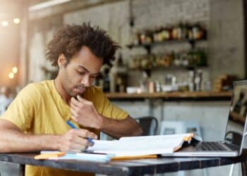 Homem estudando com laptop e fazendo anotações em mesa de café estabelecendo rotina de estudos para concurso