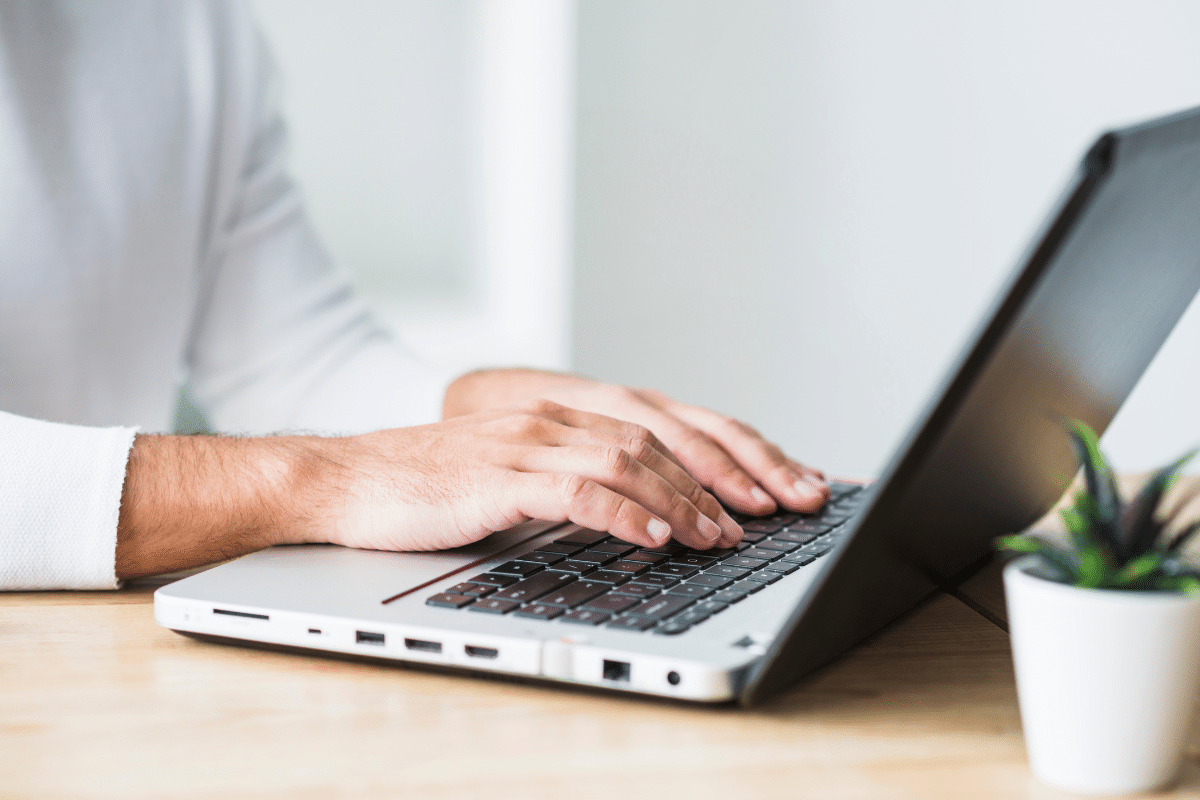 Duas mãos masculinas digitando no teclado de notebook prata sobre mesa de madeira clara