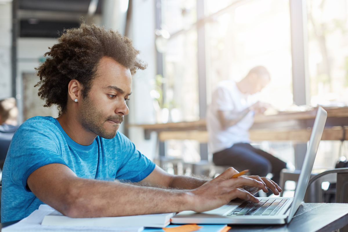 Concurso Prefeitura Homem jovem com cabelo afro concentrado trabalhando em laptop com caderno aberto em ambiente claro e moderno