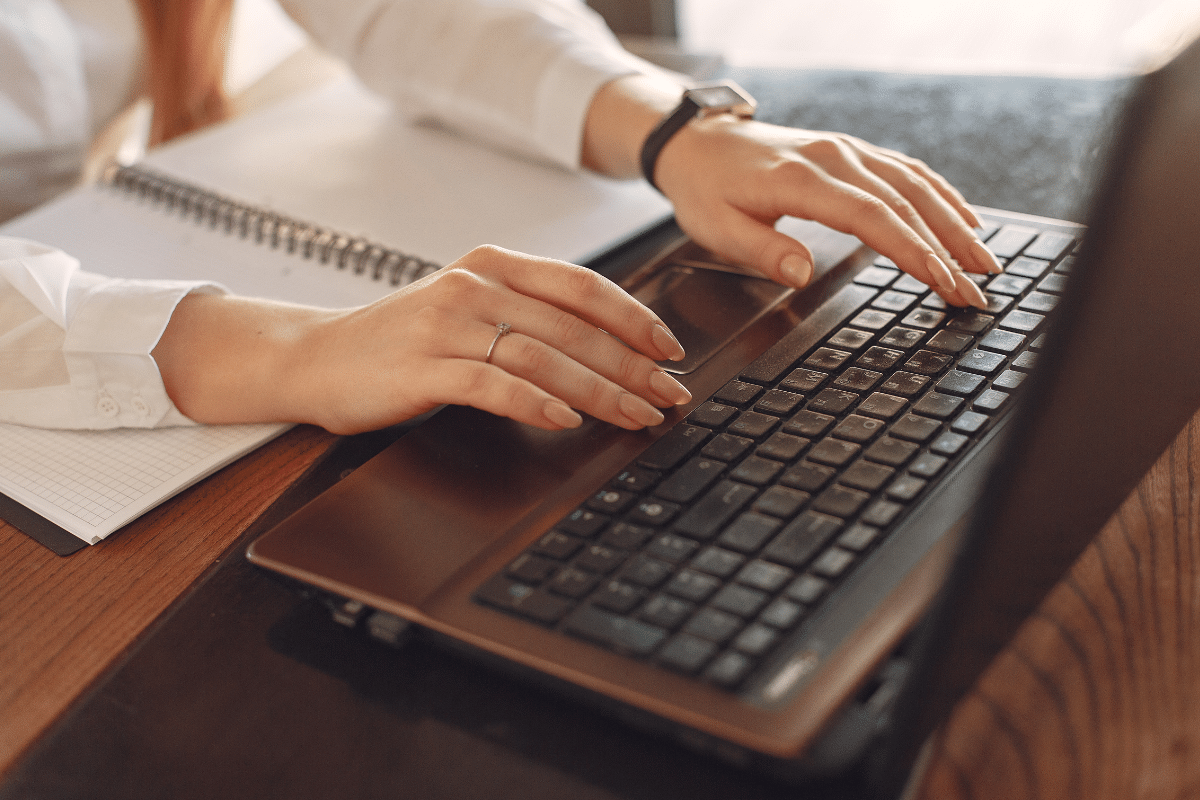 Mãos femininas digitando em teclado de notebook com caderno ao lado em mesa de madeira