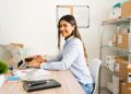 Mulher sorridente trabalhando no computador em um escritório, com mesa organizada e ambiente de trabalho moderno.