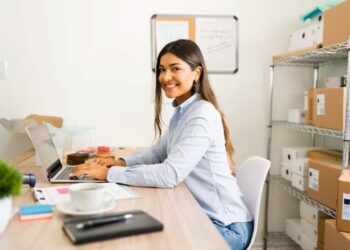 Mulher sorridente trabalhando no computador em um escritório, com mesa organizada e ambiente de trabalho moderno.