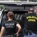 Dois policiais civis de costas com uniformes pretos próximos a viatura em operação policial