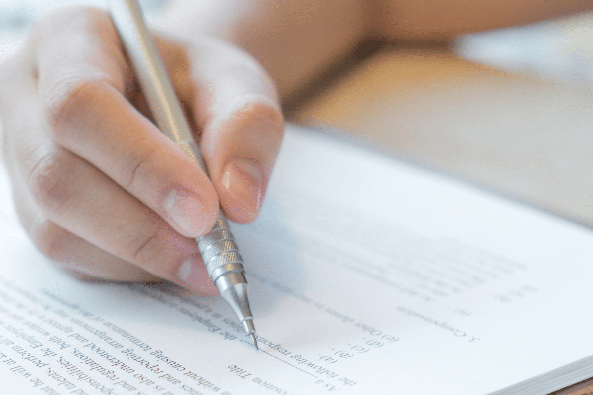 Mão segurando caneta prateada e escrevendo em papel com texto impresso