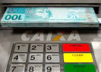 Notas de R$ 100 sendo retiradas de um caixa eletrônico da CAIXA, com a tela do terminal visível.