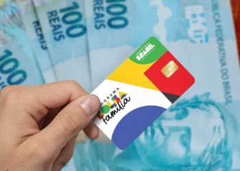 Mão segurando cartão do programa Bolsa Família com várias notas de 100 reais ao fundo