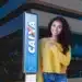 Mulher sorridente aponta para letreiro da Caixa Econômica Federal, em frente a uma agência.