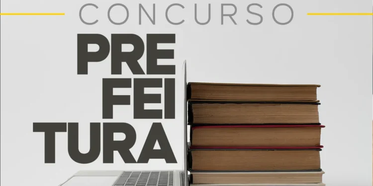 Computador aberto ao lado de pilha de livros, representando estudo para concurso de prefeitura