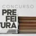 Computador aberto ao lado de pilha de livros, representando estudo para concurso de prefeitura