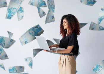 Mulher digitando em laptop com notas de 100 reais voando ao redor, representando pagamento extra.