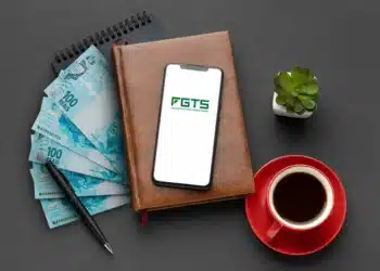 "Notas de 100 reais e celular com logo do FGTS sobre uma mesa com café, caderno e planta. Imagem relacionada ao saque-aniversário do FGTS.