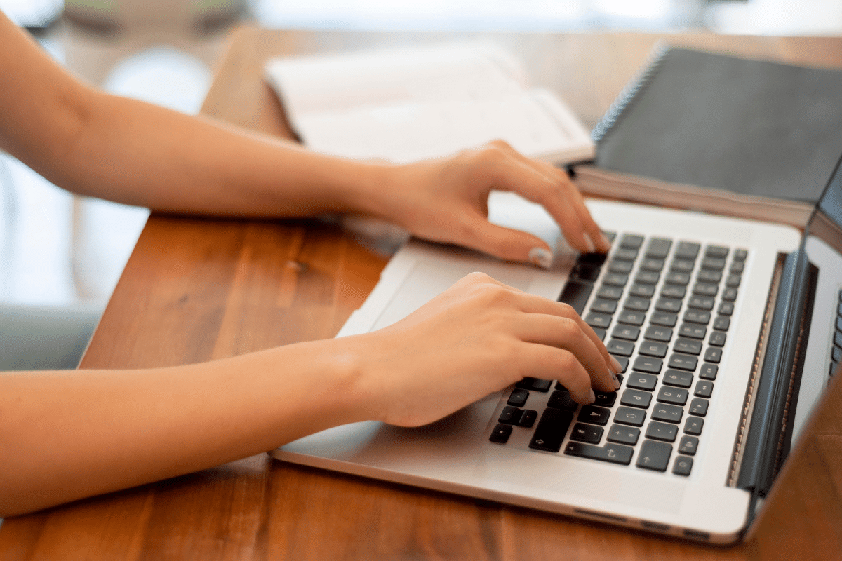 Duas mãos femininas digitando em teclado de notebook preto e prata sobre mesa de madeira