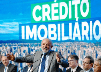 Homem de terno falando em microfone com fundo de tela gigante escrito Crédito Imobiliário e pessoas sentadas ao fundo.