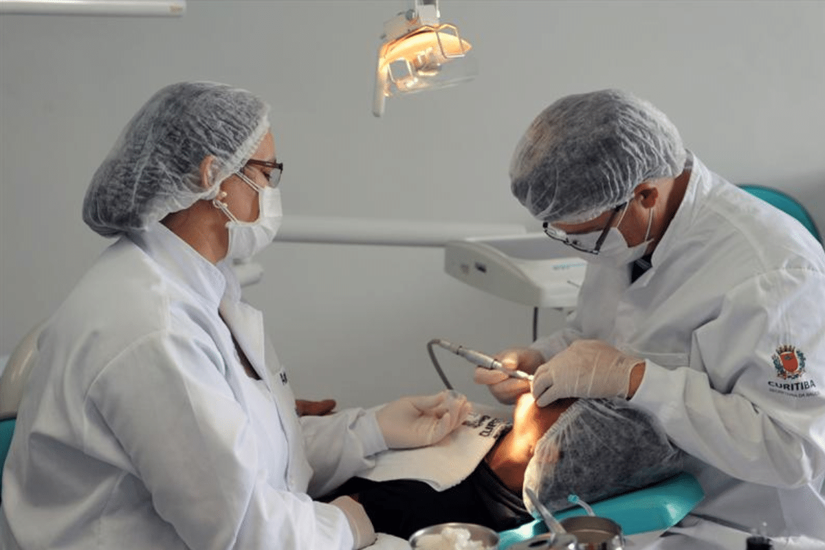 Profissionais de odontologia usando jalecos e máscaras atendem paciente em cadeira odontológica em clínica