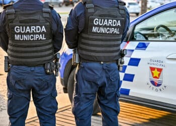 Dois guardas municipais de costas em uniforme azul com colete preto e viatura oficial à direita em Vitória