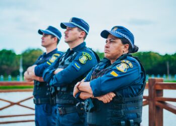 Três guardas municipais com uniforme azul, colete à prova de balas e boné, olhando concentrados com braços cruzados.