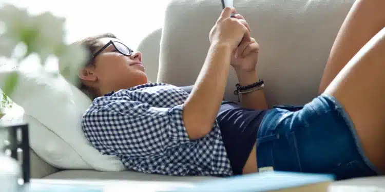 Pessoa relaxando no sofá e usando o celular, ilustrando a procrastinação.