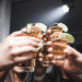 Mãos segurando copos de shot com bebida dourada e fatias de limão em ambiente de festa