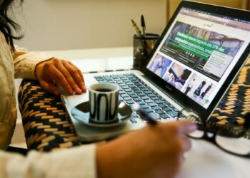 Mulher em casa em teletrabalho digitando no notebook e fazendo anotações em uma folha, com xícara de café ao lado, simbolizando a flexibilidade da carreira híbrida.
