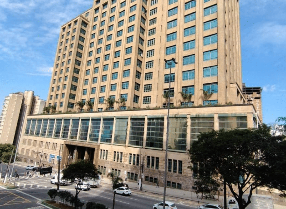 Foto do edifício da Secretaria da Fazenda do Estado de São Paulo (Sefaz SP), um prédio alto com fachada bege, janelas amplas de vidro na base, árvores no terraço, rua com veículos e pedestres em frente, sob céu azul claro.