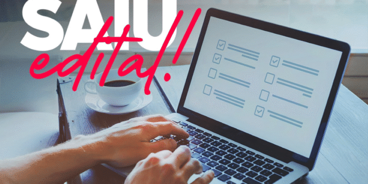 Imagem ilustrativa de mãos digitando em um laptop com tela exibindo checklist, xícara de café ao lado e texto overlay "SAIU edital!" em rosa e branco.
