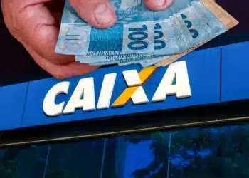 Pessoa segurando várias cédulas de 100 reais acima da fachada azul da Caixa Econômica Federal
