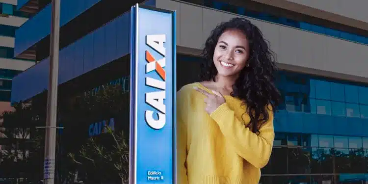 Mulher usa blusa amarela e jeans apontando para placa da Caixa em prédio moderno.