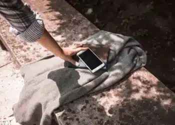 Uma mão tocando um smartphone e um caderno sobre um tecido, com sombras projetadas na superfície ao redor.