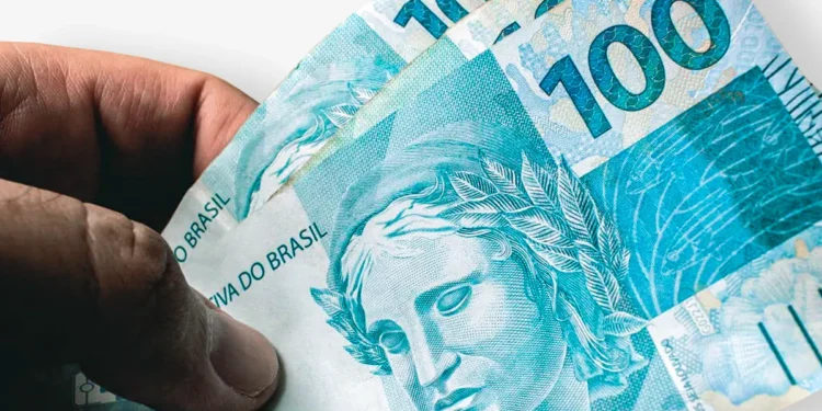 Mão de uma pessoa segurando três notas de R$ 100, totalizando R$ 300, que é o valor do cartão-alimentação.
