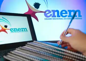 Pessoa com caneta escrevendo em cadernos com logotipo ENEM no computador e na tela maior