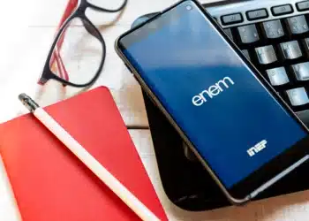 Smartphone com o aplicativo ENEM e logotipo do INEP em tela azul, ao lado de óculos de grau, lápis e um caderno de anotações vermelho, simbolizando a preparação final para a prova do Exame Nacional do Ensino Médio de 2025.