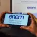 Mãos segurando celular com logo do Enem na tela e computador ao fundo.